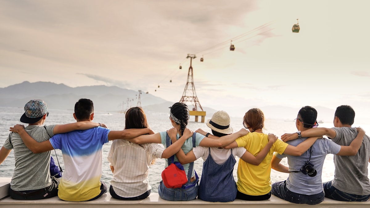 A diverse group of friends sitting together, arms around each other, looking out over a scenic waterfront
